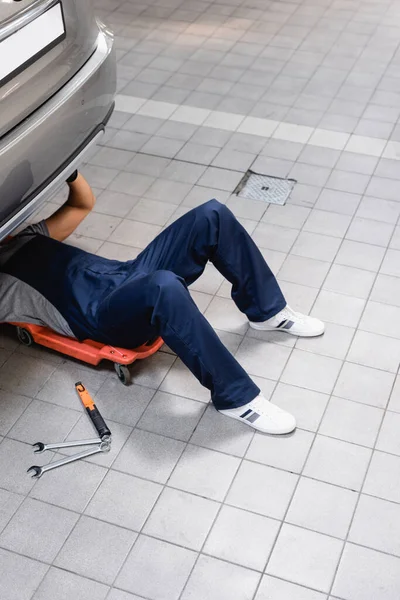 Mechanic in uniform lying under car near metallic tools — Stock Photo