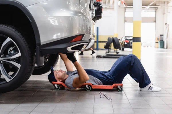 Beau mécanicien en uniforme et chapeau couché sous la voiture dans le centre de service — Photo de stock