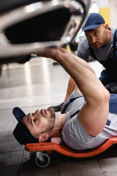 Foco seletivo do mecânico mentindo e reparando auto perto do colega de trabalho no centro de serviço — Fotografia de Stock