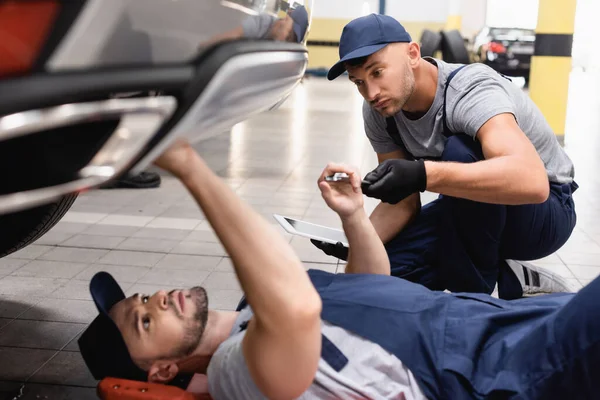 Selective focus of mechanic in uniform holding digital tablet and looking at coworker lying and repairing auto in car service — Stock Photo