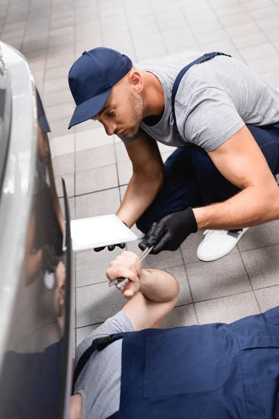 Selective focus of mechanic in uniform holding digital tablet while giving wrench to coworker lying and repairing auto — Stock Photo