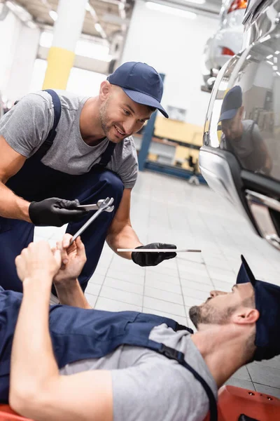 Selektiver Fokus des glücklichen Mechanikers mit digitalem Tablet und Schraubenschlüssel in der Nähe eines Mitarbeiters in einem Reparaturauto — Stockfoto