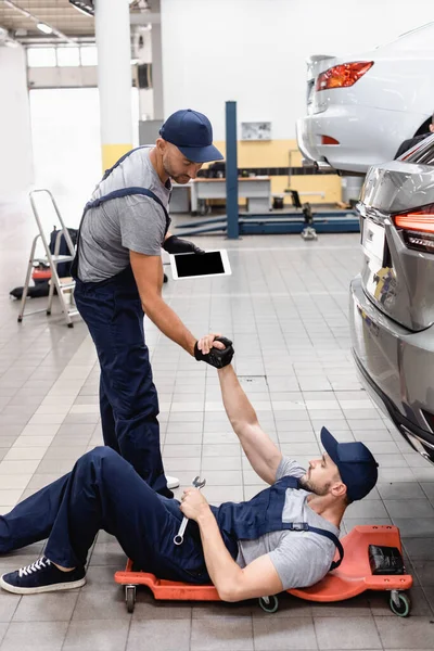 Mechanic with digital tablet holding hands with coworker in cap lying near car — Stock Photo