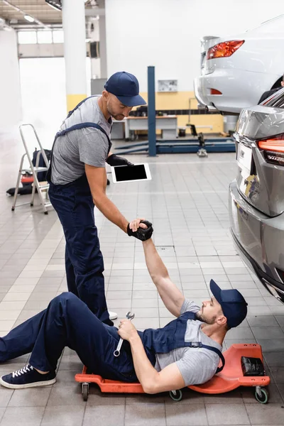 Mecánico con tableta digital cogido de la mano con su compañero de trabajo en la tapa acostado cerca del coche - foto de stock