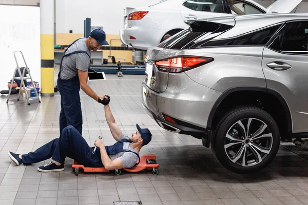 Mechanic holding hands with coworker in cap lying near cars — Stock Photo