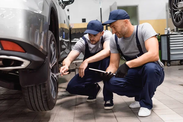 Mechanic holding digital tablet near coworker with wrench repairing car — Stock Photo