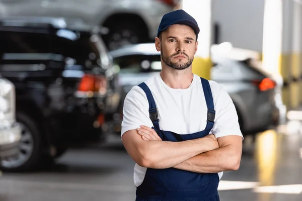 Beau mécanicien en uniforme et casquette debout avec bras croisés en service de voiture — Photo de stock