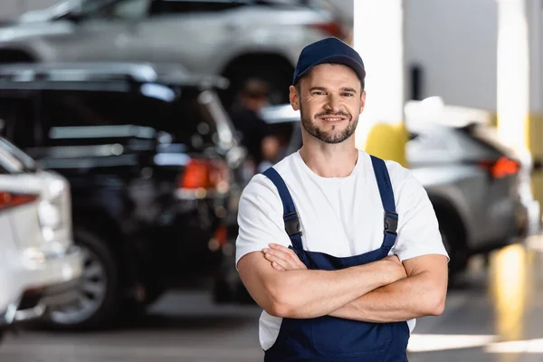 Feliz mecánico en uniforme y gorra de pie con brazos cruzados en servicio de coche — Stock Photo