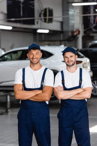 Happy mechanics in uniform and caps standing with crossed arms in car service — Stock Photo