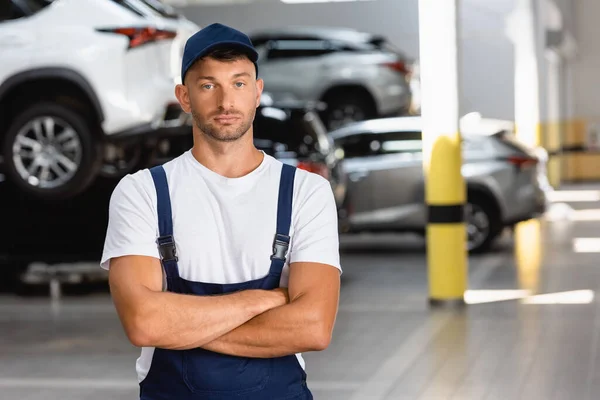Schöner Mechaniker in Uniform und Mütze, der mit verschränkten Armen in der Tankstelle steht — Stockfoto