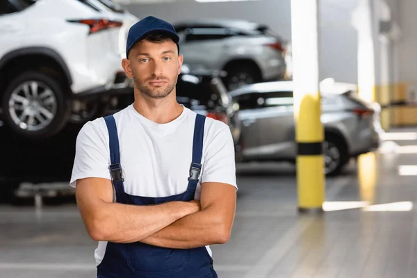 Guapo mecánico en uniforme y gorra de pie con los brazos cruzados en la estación de servicio - foto de stock