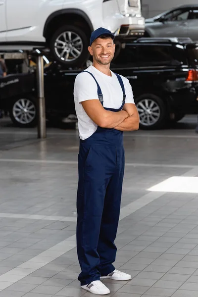 Cheerful mechanic in uniform and cap standing with crossed arms in service station — Stock Photo