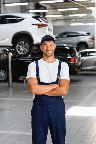 Glücklicher Mechaniker in Uniform und Mütze mit verschränkten Armen in der Tankstelle — Stockfoto
