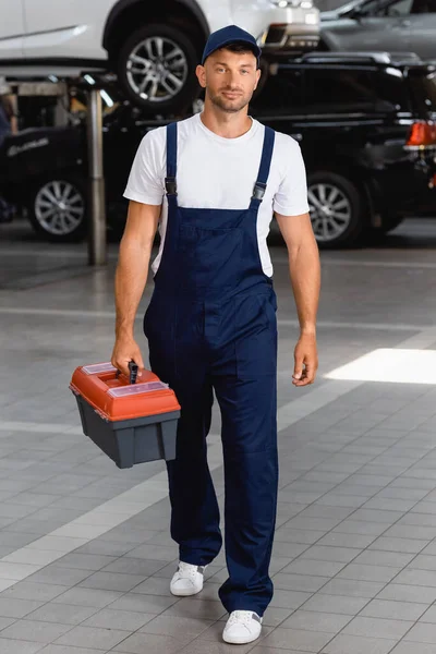 Handsome mechanic in uniform and cap holding toolbox in service station — Stock Photo