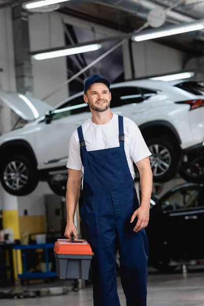 Heureux mécanicien en salopette et casquette tenant boîte à outils dans le service de voiture — Photo de stock