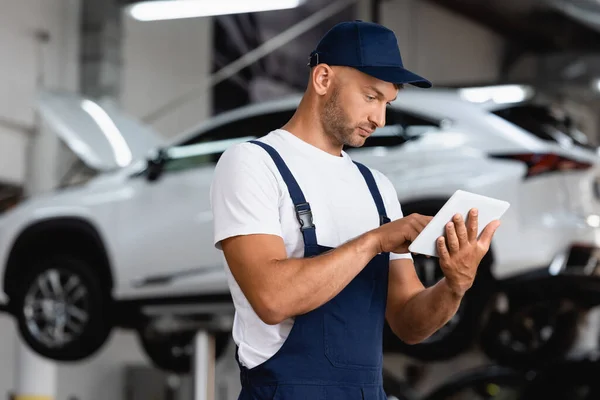 Handsome mechanic in overalls and cap using digital tablet in car service — Stock Photo