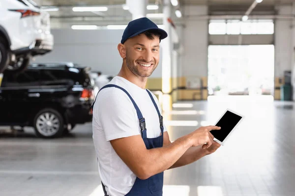 Heureux mécanicien en salopette et chapeau pointant avec le doigt à la tablette numérique avec écran blanc dans le service de voiture — Photo de stock