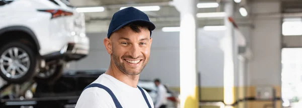 Panoramic shot of happy mechanic in cap looking at camera — Stock Photo