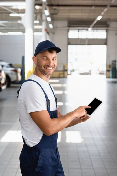 Cheerful mechanic in overalls and cap pointing with finger at digital tablet with blank screen in car service — Stock Photo