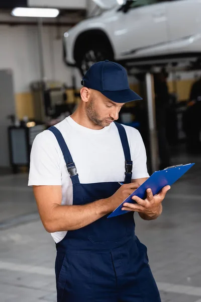 Handsome mechanic in uniform and cap writing while holding clipboard — Stock Photo