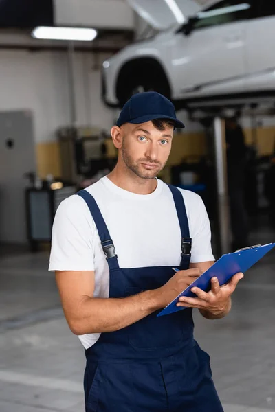Handsome mechanic in uniform and cap writing while holding clipboard and looking at camera — Stock Photo
