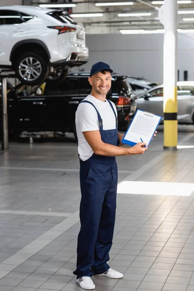 Cheerful mechanic in overalls and cap holding clipboard with contract lettering and pen near cars — Stock Photo