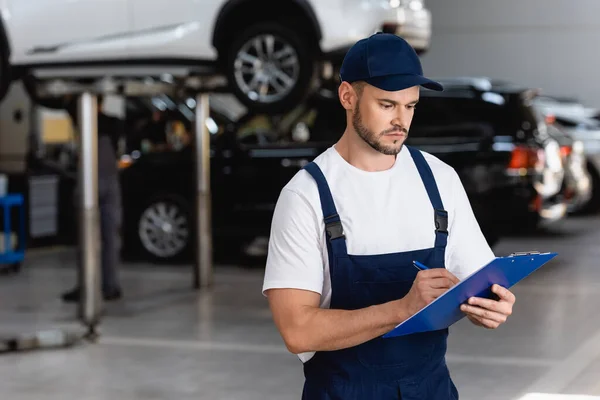 Handsome mechanic in overalls and cap writing while holding clipboard — Stock Photo