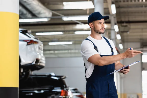 Schöner Mechaniker in Uniform und Mütze, der mit der Hand zeigt, während er Klemmbrett und Stift in der Nähe von Autos hält — Stockfoto