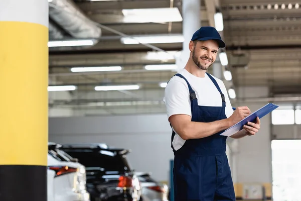 Mecánico feliz en uniforme y la escritura de la tapa mientras que sostiene el portapapeles y la pluma cerca de coches — Stock Photo