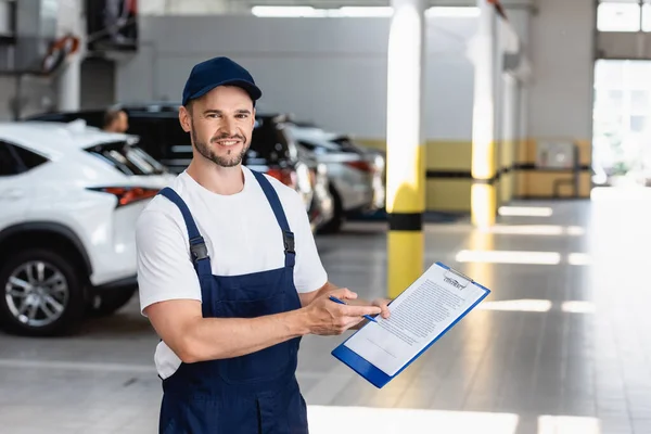 Glücklicher Mechaniker in Uniform und Mütze mit Klemmbrett mit Vertragsaufschrift und Stift in der Nähe von Autos — Stockfoto
