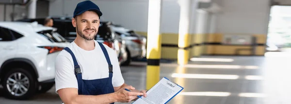 Panoramic concept of happy mechanic in uniform and cap holding clipboard with contract lettering and pen near cars — Stock Photo