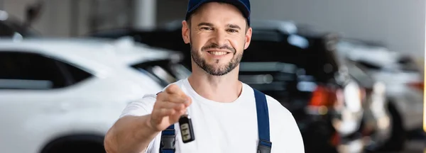 Horizontal image of cheerful mechanic in uniform and cap holding car key — Stock Photo