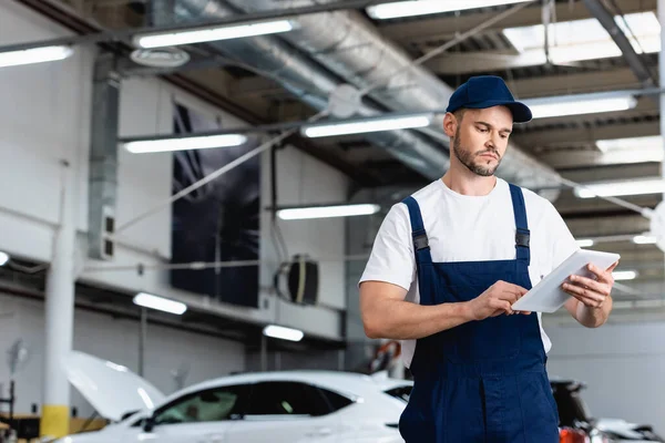 Mecânico em uniforme e tampa usando comprimido digital na oficina — Fotografia de Stock