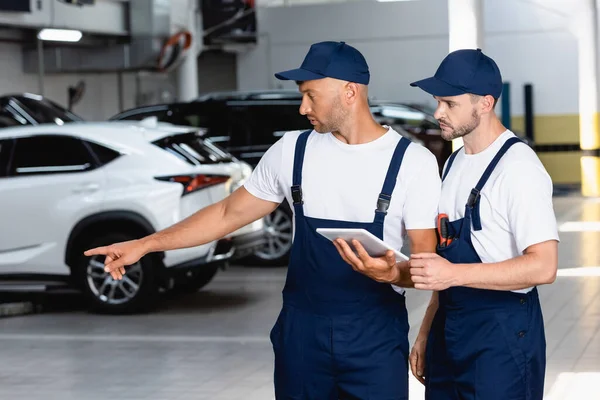 Handsome mechanic in uniform holding digital tablet and pointing with finger at car near coworker in workshop — Stock Photo