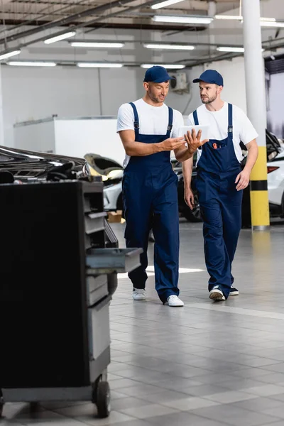 Selective focus of handsome mechanics in uniform using digital tablet and walking in workshop — Stock Photo