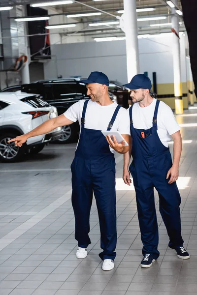Heureux mécanicien en uniforme tenant tablette numérique et pointant du doigt à la voiture près de collègue dans l'atelier — Photo de stock