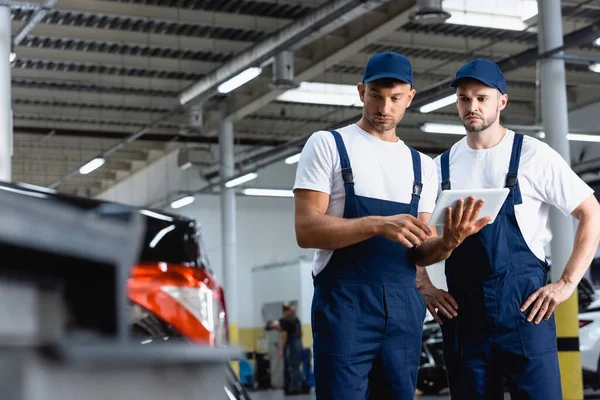 Selective focus of handsome mechanics in uniform using digital tablet and looking at car in workshop — Stock Photo