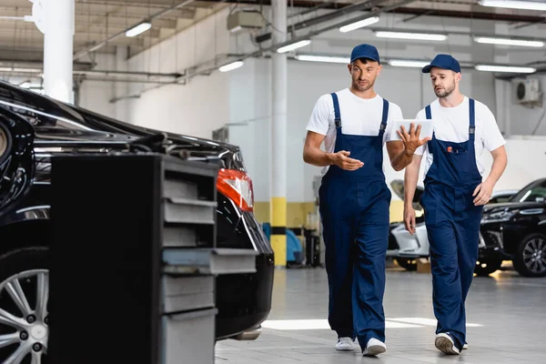 Selective focus of handsome mechanics in uniform using digital tablet and walking near cars in workshop — Stock Photo