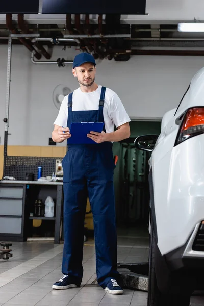 Meccanico in uniforme che tiene appunti mentre guarda l'auto — Foto stock