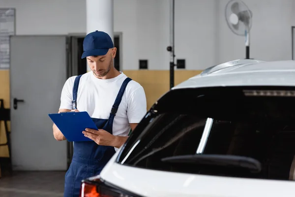 Mechaniker in Mütze hält Klemmbrett und schreibt neben Auto — Stockfoto