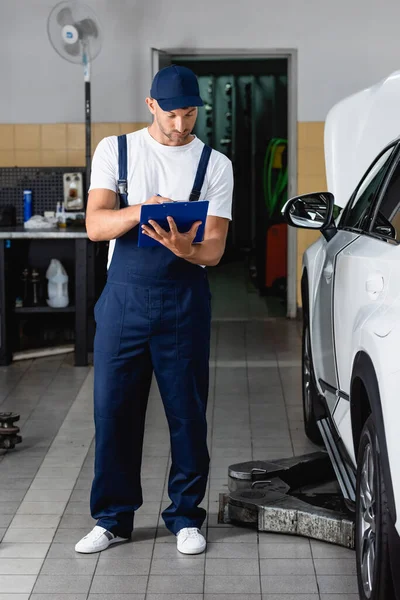 Mecánico guapo en la tapa que sostiene el portapapeles y la escritura cerca del coche - foto de stock