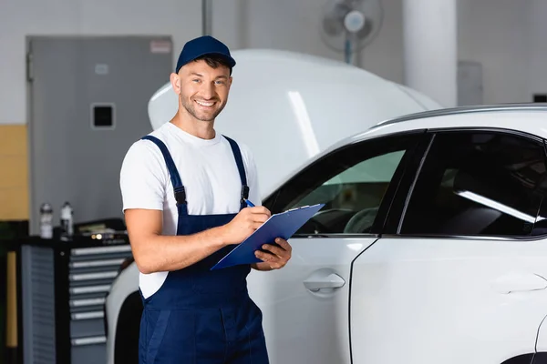 Mecánico feliz en la tapa sujetando portapapeles cerca de coche moderno - foto de stock