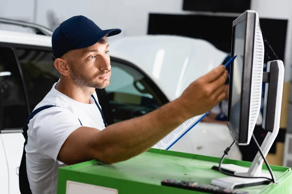 Messa a fuoco selettiva del meccanico nel cappuccio che tiene la penna vicino al monitor del computer in officina — Foto stock