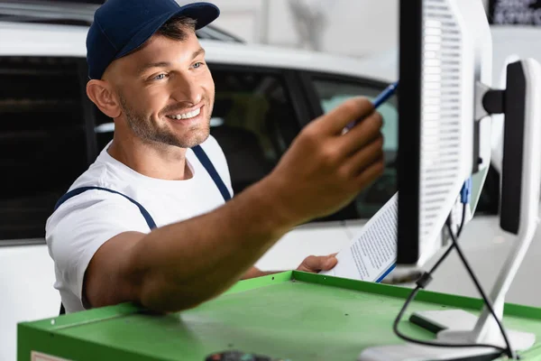 Selective focus of happy mechanic in cap holding pen near computer monitor in workshop — Stock Photo