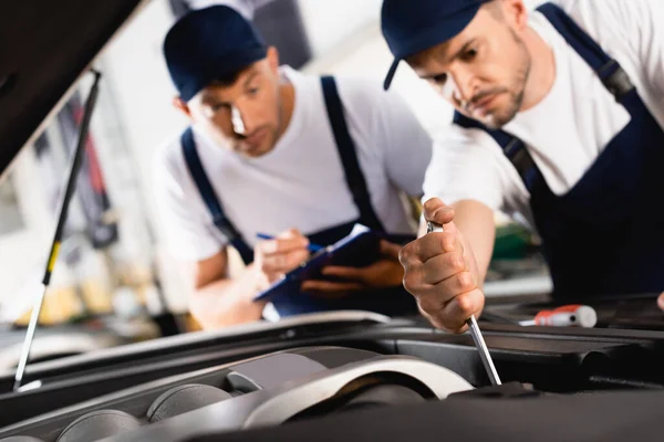 Enfoque selectivo de mecánico en la reparación de la tapa de coche cerca de compañero de trabajo con portapapeles y pluma - foto de stock
