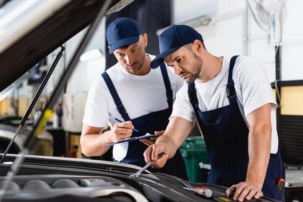 Selektiver Fokus des Mechanikers in Uniform, der sein Auto mit Klemmbrett und Stift in der Nähe des Mitarbeiters fixiert — Stockfoto