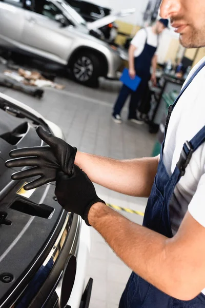 Vista recortada del mecánico en uniforme con guantes de látex cerca del coche en el taller - foto de stock