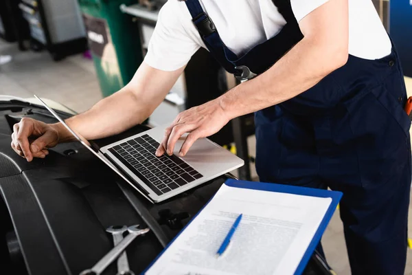 Cropped view of auto mechanic typing on laptop on hood near wrenches and clipboard at service station — Stock Photo