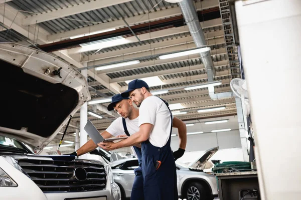 Seitenansicht des Automechanikers, der Laptop in der Nähe der offenen Motorhaube des Autos mit einem Kollegen an der Tankstelle hält — Stockfoto