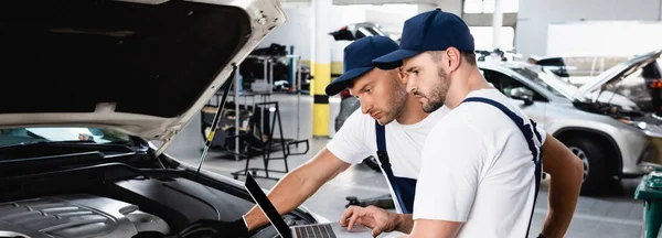 Panoramic shot of auto mechanics looking at laptop screen near car at service station — Stock Photo